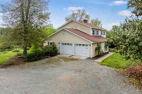 Side view of the renovated house at 1701 Harrington Road, Orange VA, showing attached two-car garage, red roof, gravel driveway, and surrounding greenery.