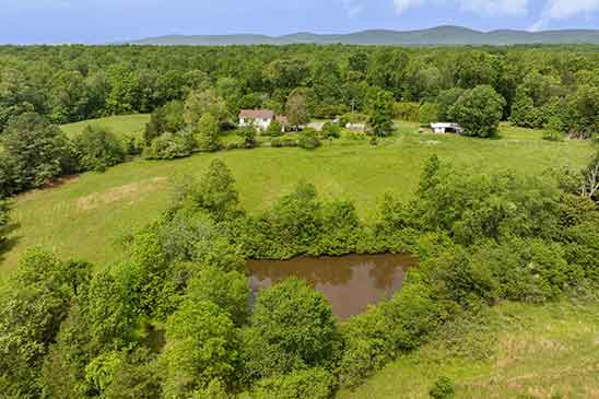 Scenic aerial view of 1701 Harrington Road in Gordonsville, VA, showcasing the renovated farmhouse, private pond, rolling pastures, and Blue Ridge Mountain backdrop.