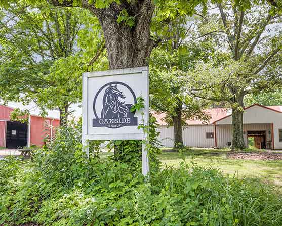 Oakside Stables sign mounted on a tree with a horse logo, surrounded by lush greenery, with red and white barn buildings visible in the background at 2205 S. Amherst Hwy, Amherst, VA 24521.