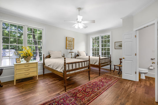 Main-level en-suite guest bedroom at 3665 Old River Trail in Powhatan, Virginia, featuring twin beds, hardwood floors, large windows, and a private bathroom. This comfortable space is ideal for guests or extended family living at Pleasant Oak.