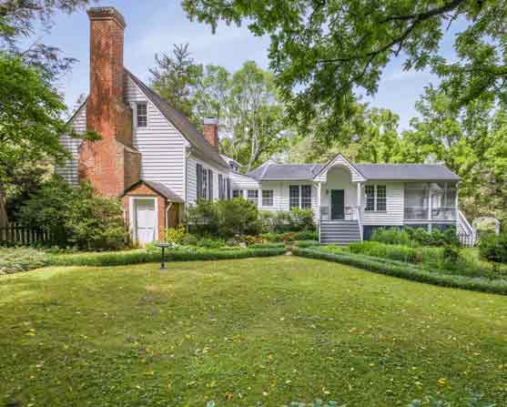 Charming colonial-era home with white siding and tall brick chimneys, featuring a graceful addition, landscaped gardens, and a formal lawn framed by boxwood hedges under a canopy of mature trees.