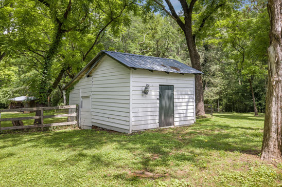 Historic smokehouse at 3665 Old River Trail in Powhatan, Virginia, with wood siding, tin roof, and fenced backdrop. A contributing structure to the 18th-century character of Pleasant Oak, reflecting early American homestead life.