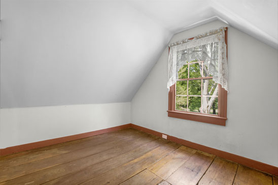Small upstairs room at 3665 Old River Trail in Powhatan, Virginia, with original wide-plank floors, angled ceiling, and a vintage window overlooking the grounds. A quaint historic space in the original section of Pleasant Oak.