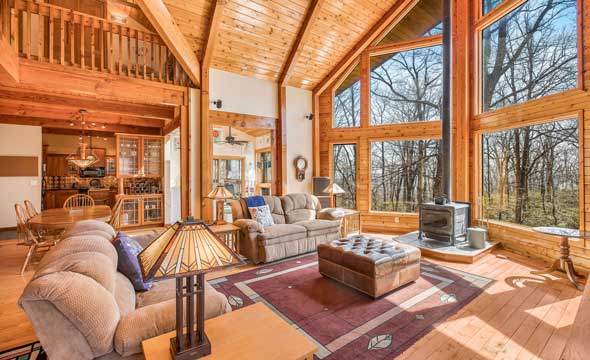 Bright and inviting living room with vaulted wood ceilings, expansive floor-to-ceiling windows, a wood stove, plush seating, and an open-concept layout leading to the dining area and kitchen.